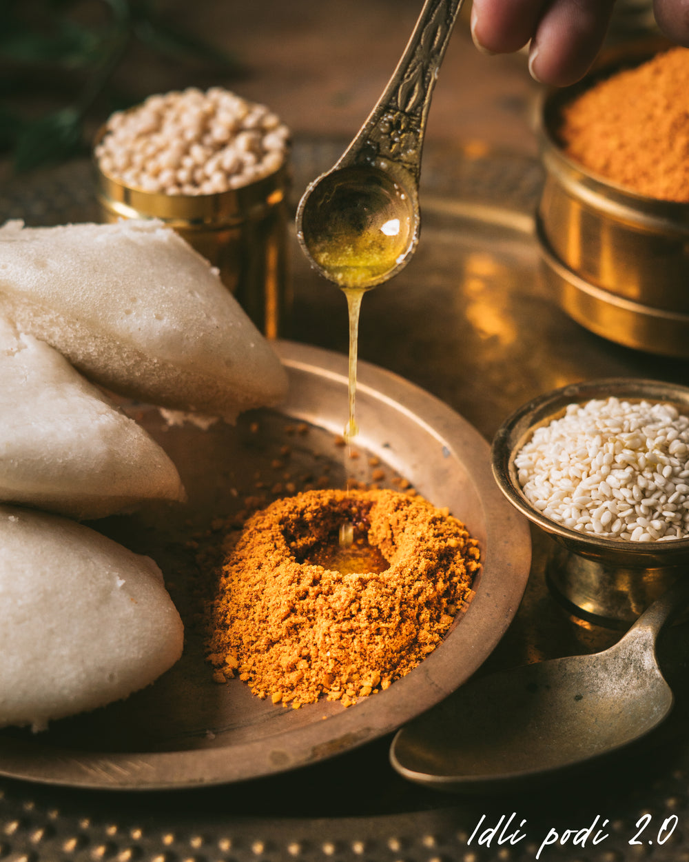 Idli with chutney and spices on a metal tray, with a hand holding a spoon.